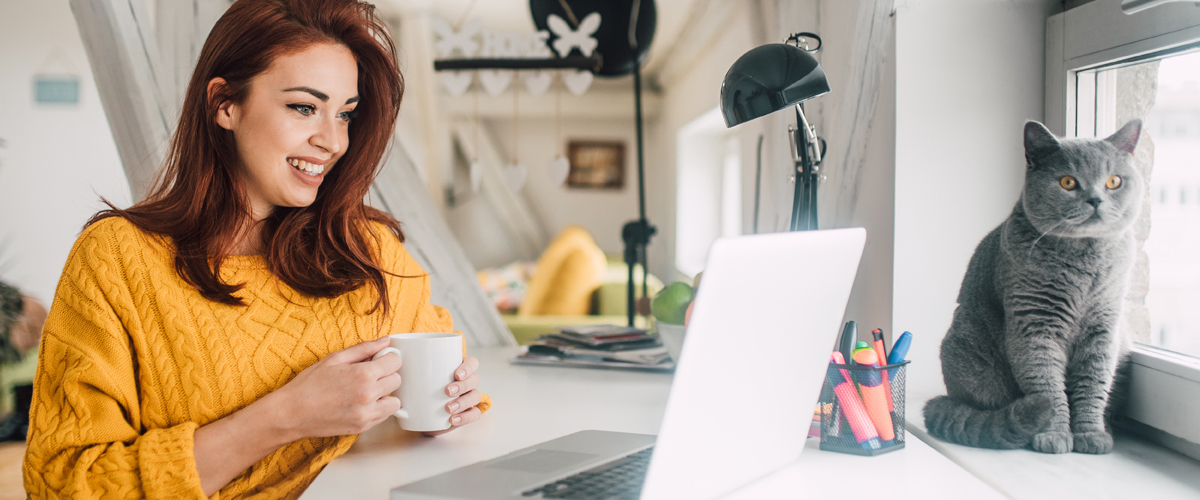 Young woman sipping from a mug while looking at her laptop. Her cat is beside her looking out the nearby window.
