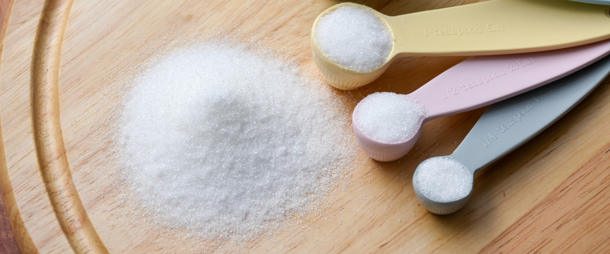 A small pile of granulated white sugar sits on a wooden surface next to several measuring spoons filled with sugar.